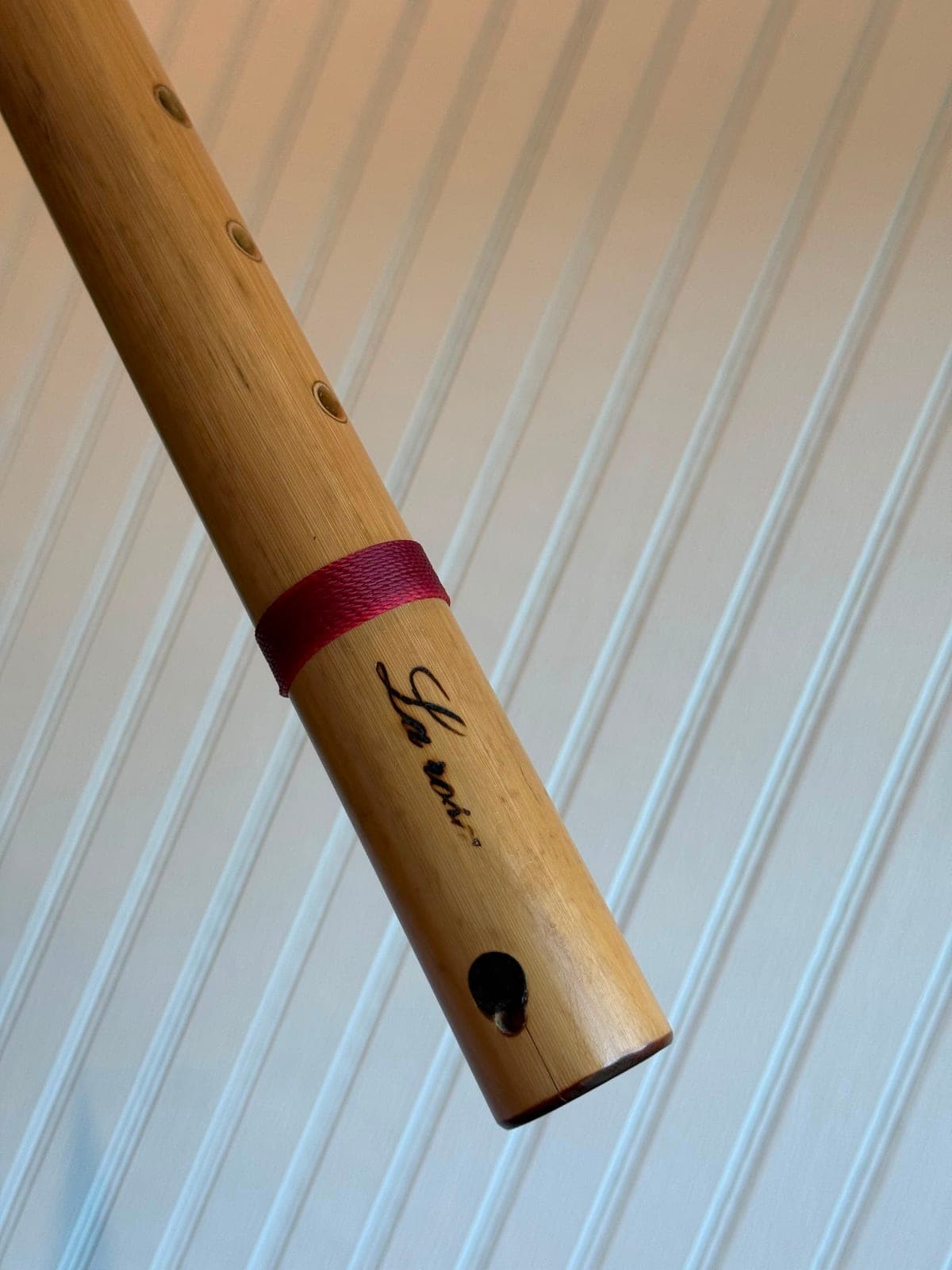The finished bamboo flute wrapped with a red waxed cord and marked with a small carved bird, against a pale beadboard wall