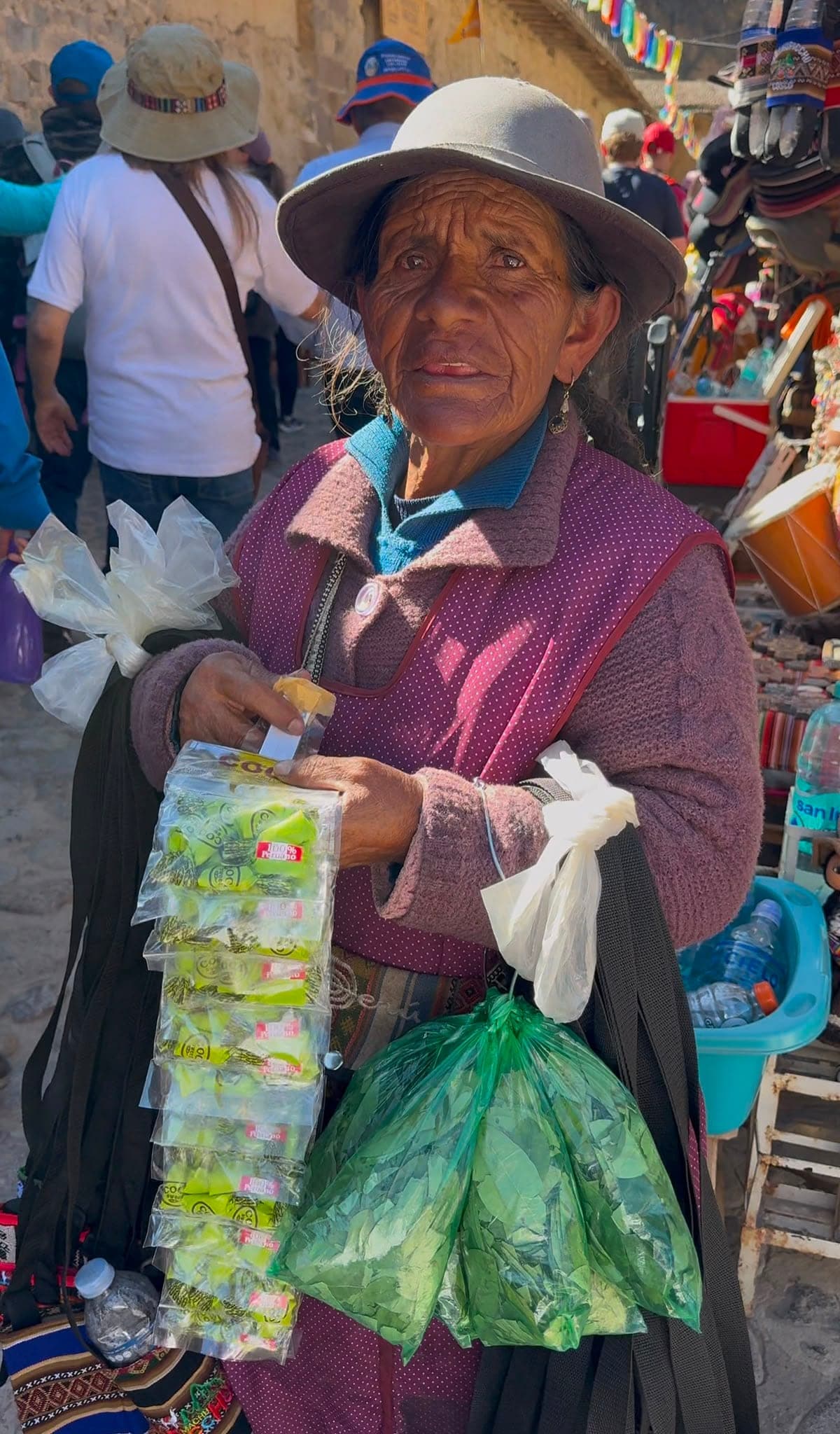 An elderly Peruvian woman in a hat holding out strips of coca candy and a bag of goods to sell in the busy market