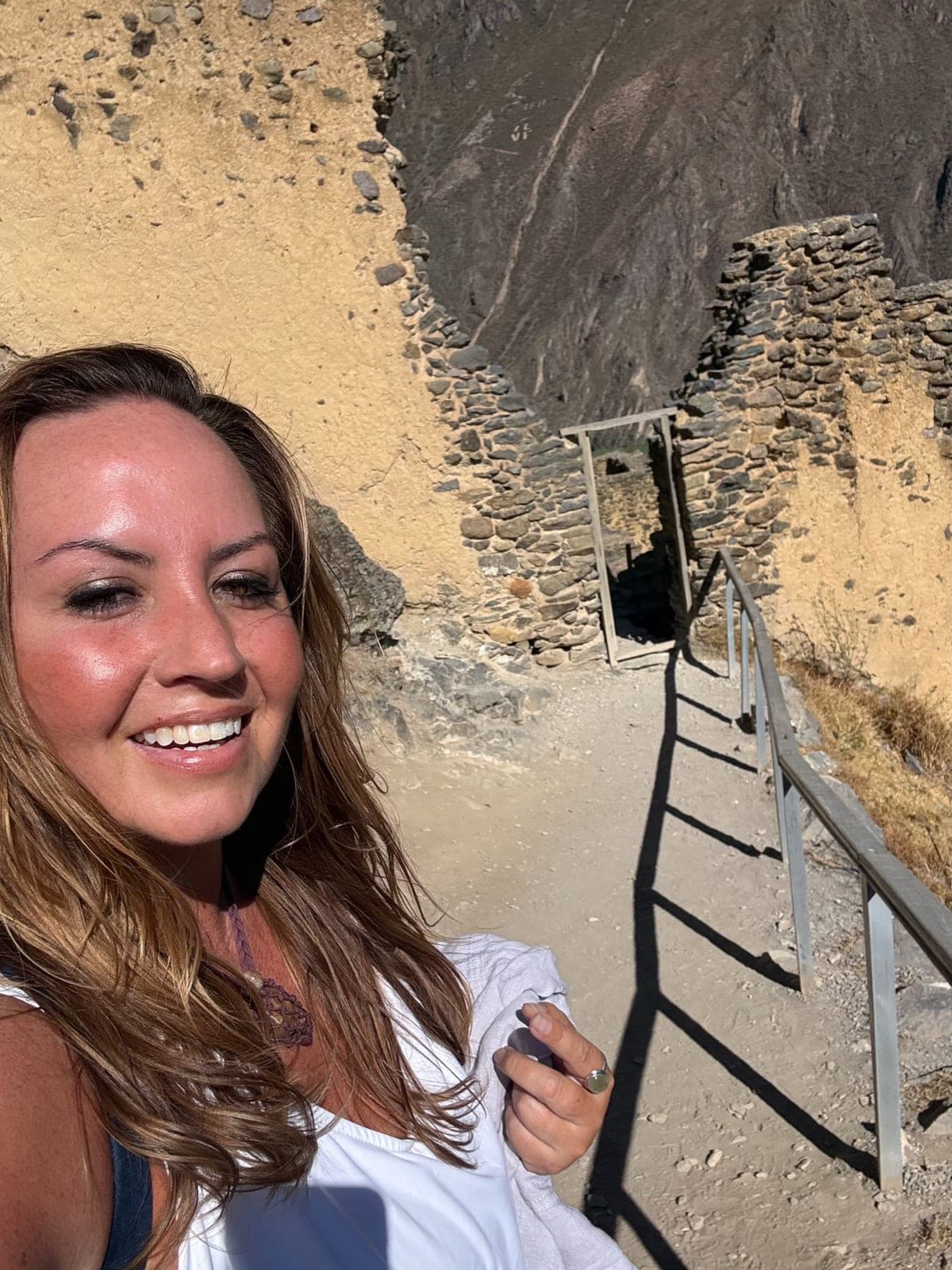 Sarah smiling in a white top on the stone steps of Ollantaytambo, ruins and a railed path climbing behind her