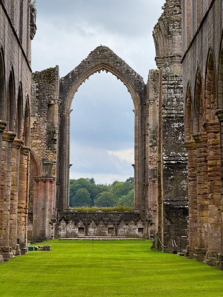 The roofless nave of a ruined stone abbey, a tall pointed arch standing open to the sky above cropped grass