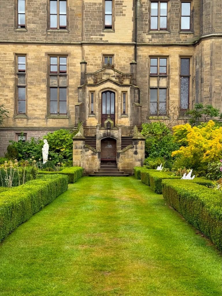 A clipped grass path between low box hedges running straight to the arched stone porch of a Gothic revival house