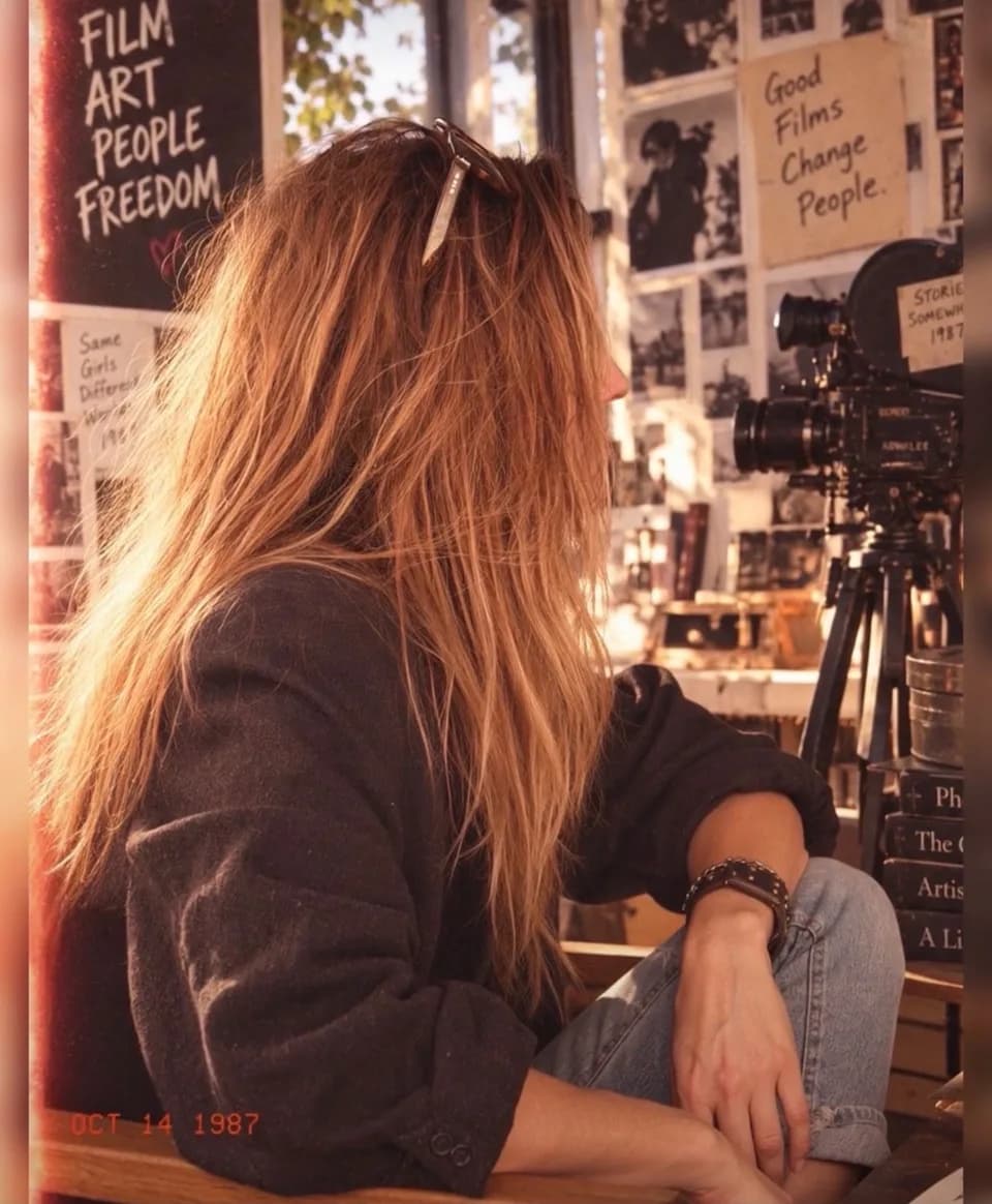 A woman with long sun-lit hair seen in profile, watching an old film camera on a tripod in a room hung with black and white prints