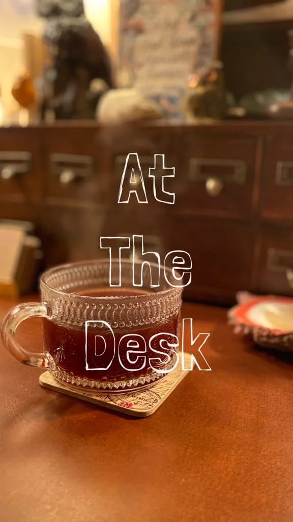 A glass mug of tea steaming on a leather desk beside a wooden card-catalogue cabinet, the words At The Desk written across it