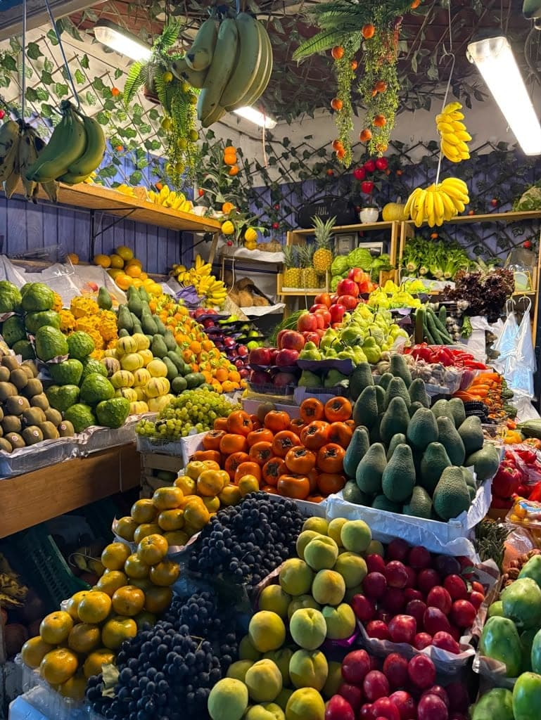 A market stall banked with fruit and vegetables: pyramids of persimmons, avocados, grapes and apples under strip lights, with bananas hanging from the ceiling