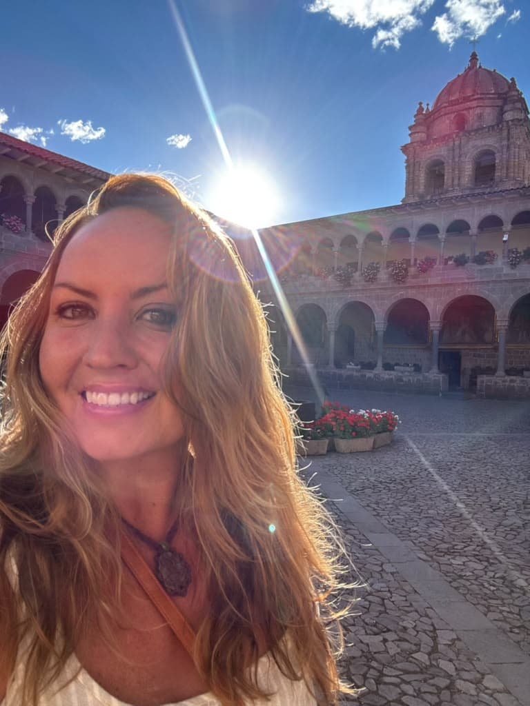 Sarah smiling in bright sunlight, an arcaded colonial courtyard and a bell tower behind her